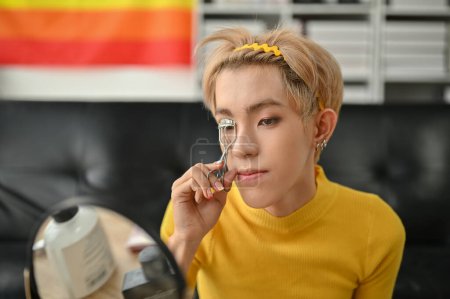 An attractive young Asian gay man is looking at the mirror and curling his eyelashes with a curler while doing his makeup at home. LGBTQ+ and beauty lifestyle concept