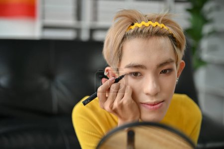 An attractive young Asian gay man applies liquid eyeliner to create a winged look while doing his makeup at home. LGBTQ+ and beauty lifestyle concepts