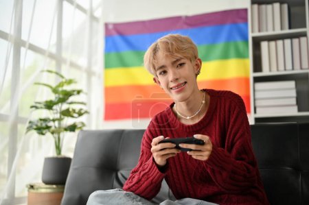 Ein attraktiver und lächelnder junger asiatischer Schwuler sitzt mit seinem Smartphone in der Hand auf einem Sofa und entspannt sich in seinem Wohnzimmer.