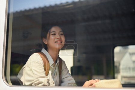 Téléchargez les photos : Une jolie et heureuse jeune voyageuse asiatique ou une routeuse s'assoit sur son siège dans un train, voyageant seule quelque part. Concept vacances d'été - en image libre de droit