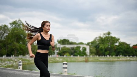 Photo for A sporty and determined Asian woman in sportswear is running along the street around the public park. Healthy lifestyle concept - Royalty Free Image