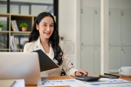 Téléchargez les photos : Une femme d'affaires asiatique millénaire attrayante ou une comptable féminine est assise à son bureau avec des rapports d'entreprise et son ordinateur portable. - en image libre de droit