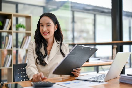 Téléchargez les photos : Une femme d'affaires asiatique millénaire charmante et souriante ou une gestionnaire féminine en costume d'affaires s'assoit à son bureau et regarde la caméra. - en image libre de droit