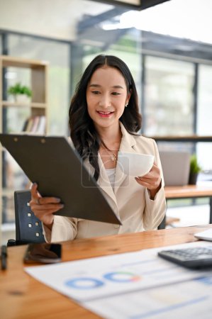 Téléchargez les photos : Portrait d'une belle femme d'affaires asiatique millénaire examinant un rapport d'entreprise et sirotant son café du matin à son bureau dans le bureau. - en image libre de droit