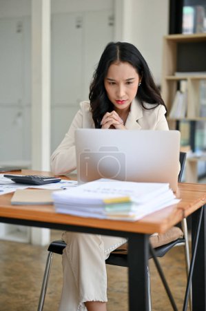 Téléchargez les photos : Un portrait d'une femme d'affaires asiatique millénaire réfléchie se concentre sur ses tâches sur son ordinateur portable, en regardant son ordinateur portable avec un visage sérieux, et en travaillant à son bureau dans le bureau. - en image libre de droit
