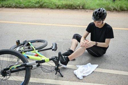 Foto de Un joven ciclista asiático herido en ropa deportiva y un casco de bicicleta se cayeron de la bicicleta mientras pedaleaba a lo largo de carreteras rurales. dolor de rodilla, hemorragia de rodilla, rodilla lesionada, accidente deportivo, lesión en bicicleta - Imagen libre de derechos
