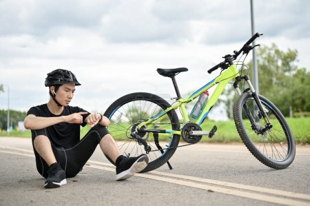 Téléchargez les photos : Un jeune cycliste asiatique fatigué assis dans la rue avec son vélo et vérifiant ses calories brûlées et chevauchant des kilomètres sur sa smartwatch. - en image libre de droit
