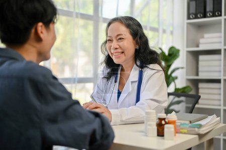 Foto de Una médica asiática anciana, cariñosa y sonriente, que sostiene las manos de una paciente para consolarla y tranquilizarla durante el chequeo médico en un hospital.. - Imagen libre de derechos