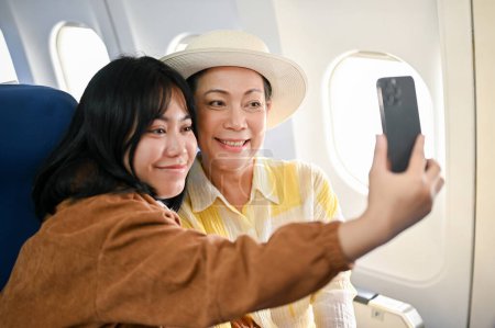 Téléchargez les photos : Une heureuse grand-mère asiatique prend des photos avec un smartphone avec sa petite-fille pendant le vol pour leurs vacances d'été ensemble. Voyage, voyage d'été, avion, mode de vie - en image libre de droit