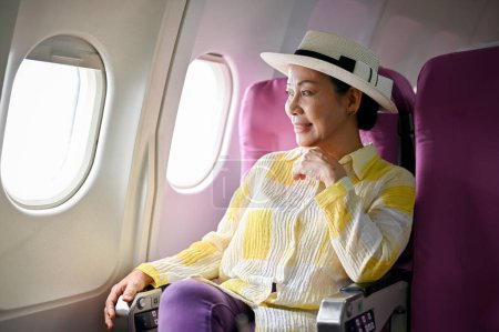Photo for An attractive and happy senior Asian female passenger is looking at the view outside of the plane from her window seat, traveling for her summer vacation. - Royalty Free Image