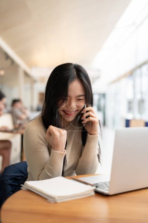 A portrait of a cheerful young Asian woman rejoicing after receiving good news over the phone while working remotely at a coffee shop co-working space.