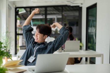 Photo for A carefree and happy young Asian businessman is leaning back in his chair and stretching his arms after finishing work while sitting at his desk in the office. - Royalty Free Image
