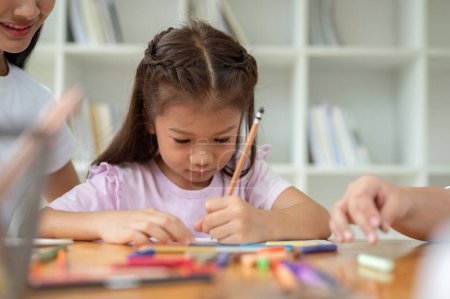 An adorable Asian elementary school girl is focusing on studying, writing, or drawing something on paper in the classroom with a teacher. Kids education concept