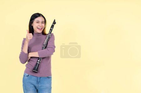 Foto de Una hermosa y talentosa joven asiática sosteniendo un clarinete y mostrando su pulgar hacia arriba, de pie sobre un fondo amarillo aislado. músico, artista, instrumentos de jazz - Imagen libre de derechos