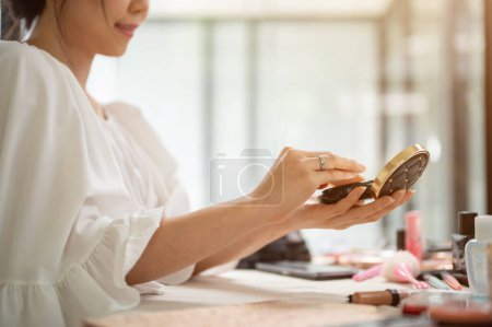 Photo for Close-up image of a beautiful Asian woman holding a compact powder or cushion, using pressed powder, doing make-up at home. rear view - Royalty Free Image