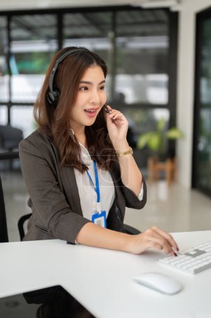 A beautiful Asian female call centre operator with a wireless headset is answering incoming calls from customers, working in a call centre customer support agency.