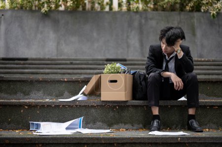 Przygnębiony, zdenerwowany i smutny młody azjatycki bezrobotny biznesmen siedzi na schodach przed biurem korporacyjnym z pudełkiem swoich osobistych rzeczy po stracie pracy..