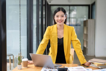 Photo for A confident and attractive millennial Asian businesswoman or female executive manager in a yellow suit stands at her office desk in her modern office. - Royalty Free Image