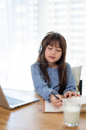 Photo for A cute young Asian girl doing homework or studying online at a table at home. Kid and education concepts - Royalty Free Image