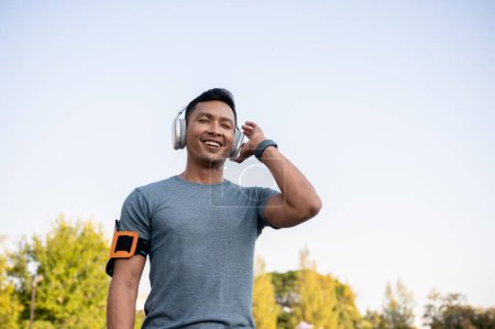 Photo for A happy and fresh Asian man in sportswear is enjoying listening to music on his wireless headphone while running in a park in the morning. - Royalty Free Image