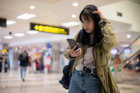 Téléchargez les photos : Une jeune voyageuse asiatique en solo confuse lit ses informations de vol sur son téléphone avec un visage sérieux et douteux, faisant face à un problème d'auto-enregistrement ou manquant son vol à l'aéroport. - en image libre de droit