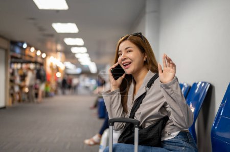 Téléchargez les photos : Une belle voyageuse asiatique parle au téléphone avec son amie alors qu'elle est assise à un siège d'attente dans le terminal de l'aéroport. - en image libre de droit