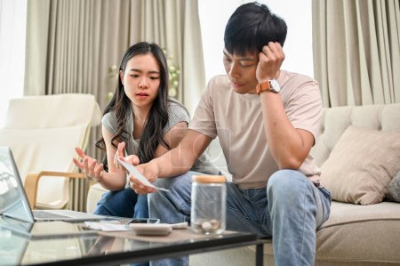 An unhappy and stressed young Asian couple is having a dispute over their household expenses and finances, arguing about high domestic bills on a sofa in the living room.
