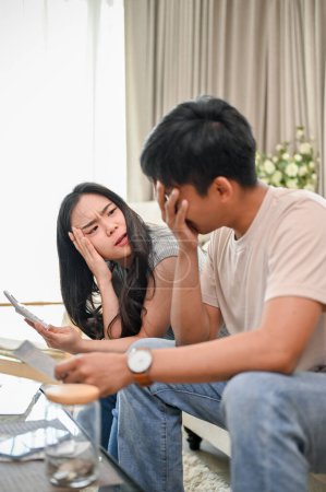An unhappy and stressed young Asian couple is having a dispute over their household expenses and finances, arguing about high domestic bills on a sofa in the living room.