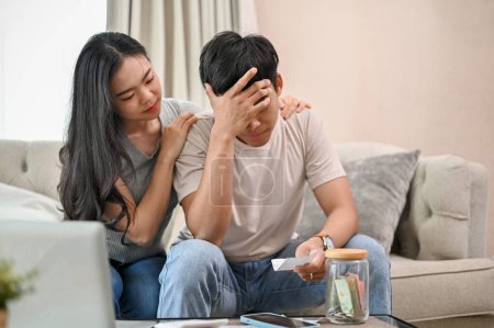 A caring young Asian woman is supporting and encouraging her stressed spouse on the sofa. The young Asian couple is worried about their household expenses and high electricity bills.