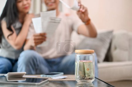 A close-up image of a jar of coins on a coffee table with a blurred background of a stressed couple worrying about their household bills and planning their finances. savings, budget, income