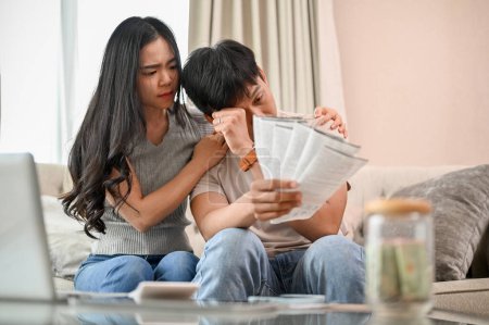 A stressed young married couple is sitting on a sofa in the living room, concerned about their invoices and household expenses. financial and couple concepts