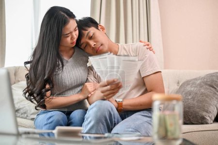 A stressed young married couple is sitting on a sofa in the living room, concerned about their invoices and household expenses. financial and couple concepts