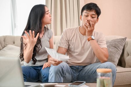 An unhappy and stressed Asian wife is arguing with her husband about unexpected expenses and high domestic bills on the sofa in the living room. spouses quarreling at home. family bankruptcy