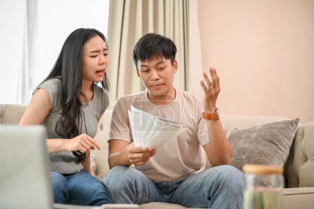 Unhappy, stressed Asian spouses are sitting on a couch, arguing about unexpected high household bills and credit card invoices. family bankruptcy, debt, lack of finances