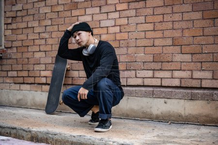 Ein cooler, stylischer asiatischer Skater in modischer Kleidung sitzt mit seinem Skateboard auf der Straße. Hipster, Breakdancer, B-Boy