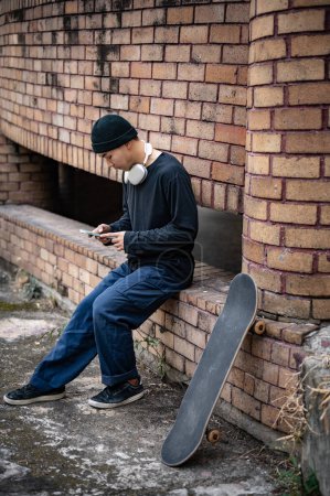 A cool, hipster Asian man in fashionable clothes with his skateboard is sitting at an unknown abandoned building in the city, using his smartphone.