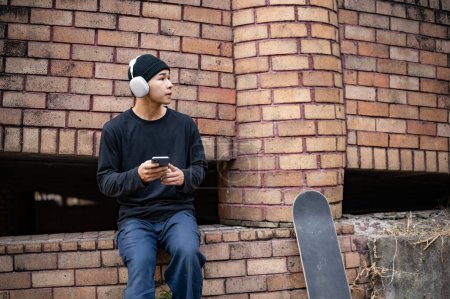 Ein cooler asiatischer Skater nutzt sein Smartphone und hört Musik über Kopfhörer, während er in einem unbekannten, verlassenen Gebäude in der Stadt sitzt.