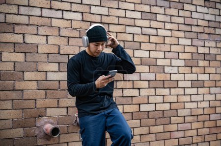 Un hombre asiático fresco y elegante con ropa de moda está usando su teléfono inteligente y escuchando música en sus auriculares mientras está de pie junto a una pared de ladrillo en la calle.