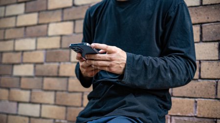 A cropped image of a cool, stylish Asian man in fashionable clothes is using his smartphone while standing by a brick wall on the street. people and wireless technology concepts