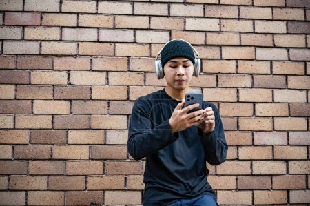 A cool, stylish Asian man in fashionable clothes is using his smartphone and listening to music on his headphones while standing by a brick wall on the street.