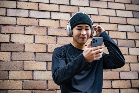 Ein fröhlicher, cooler asiatischer Mann in modischer Kleidung benutzt sein Smartphone und hört auf der Straße Musik über Kopfhörer, steht an einer Ziegelmauer.