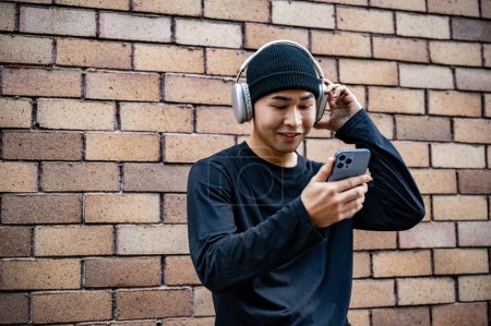 A happy, cool Asian man in fashionable clothes is using his smartphone and listening to music on his headphones on the street, standing by a brick wall.