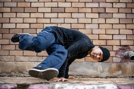 Ein professioneller, cooler asiatischer männlicher Hip-Hop-Breakdancer tanzt auf der Straße in der Nähe eines unbekannten, verlassenen Gebäudes in der Stadt und zeigt seine talentierten Bewegungen.
