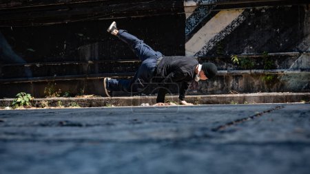 Un genial, talentoso y profesional bailarín asiático de hiphop está girando su cuerpo con las manos, bailando breakdance en la calle en una calle abandonada de la ciudad.