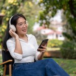A relaxed and happy Asian woman is sitting in a green backyard, listening to music on her headphones, and using her smartphone. people and lifestyle concepts