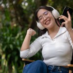 A cheerful and carefree Asian woman is enjoying the music on her headphones while sitting outdoors in her green backyard. people and lifestyle concepts