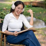 A happy and relaxed Asian woman is listening to music on her headphones, working or writing her diary while sitting in her green backyard garden.