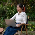 A young Asian woman is working remotely in her green backyard or at an outdoor cafe, using her laptop computer. people and technology concepts