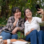 Two cheerful Asian female friends enjoy taking selfies or recording a video with a smartphone together while relaxing at an outdoor table of a cafe.