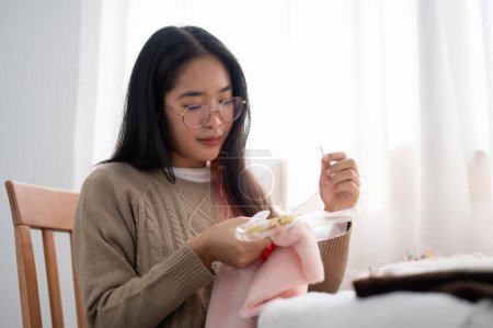 A beautiful Asian woman is focusing on hand-sewing a pattern on cloth on an embroidery frame, crafting handmade items at home. needlework, tailor, stitching
